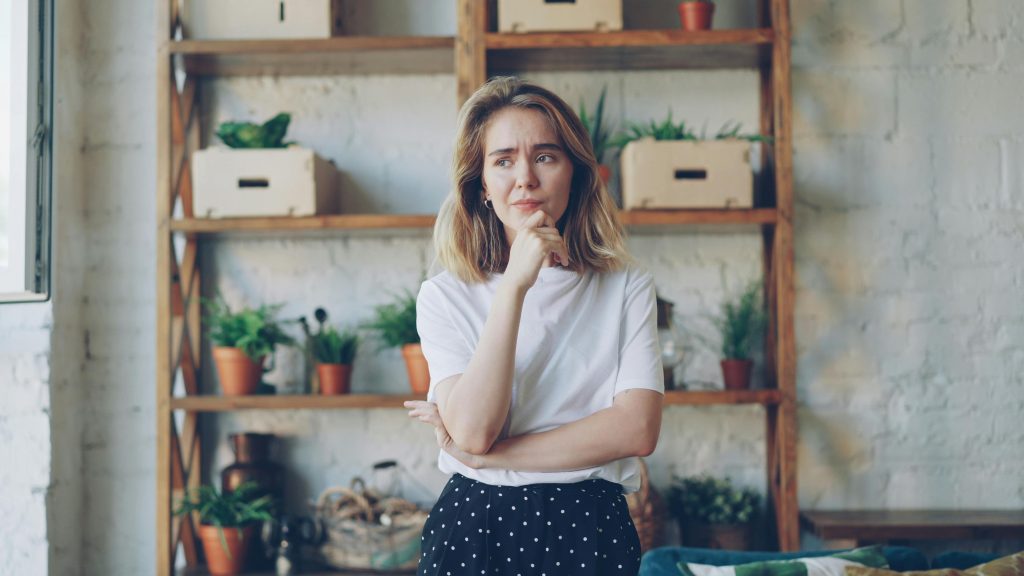 Pensive young woman in casual attire stands in a stylish room with plants.