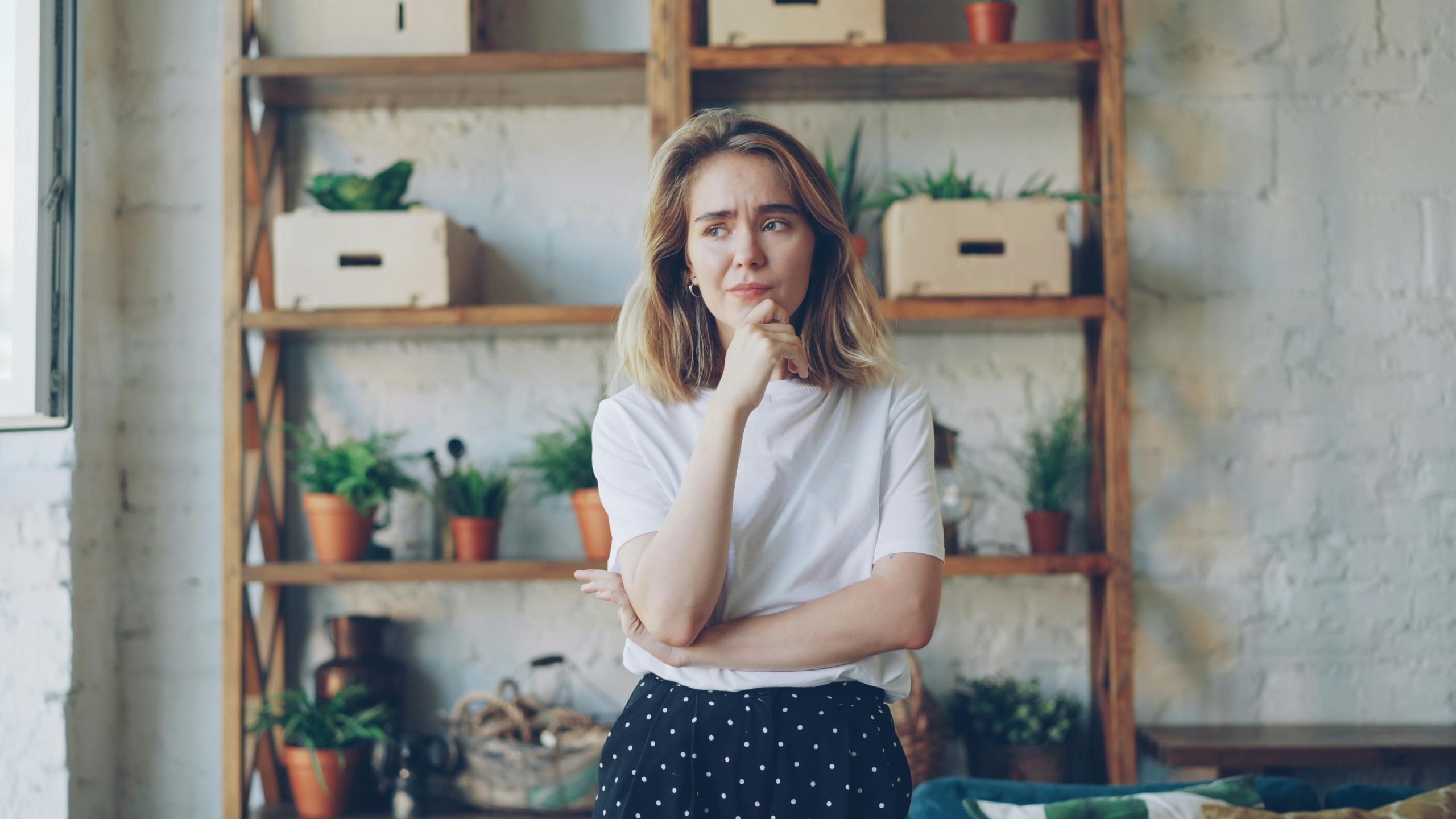 Pensive young woman in casual attire stands in a stylish room with plants.