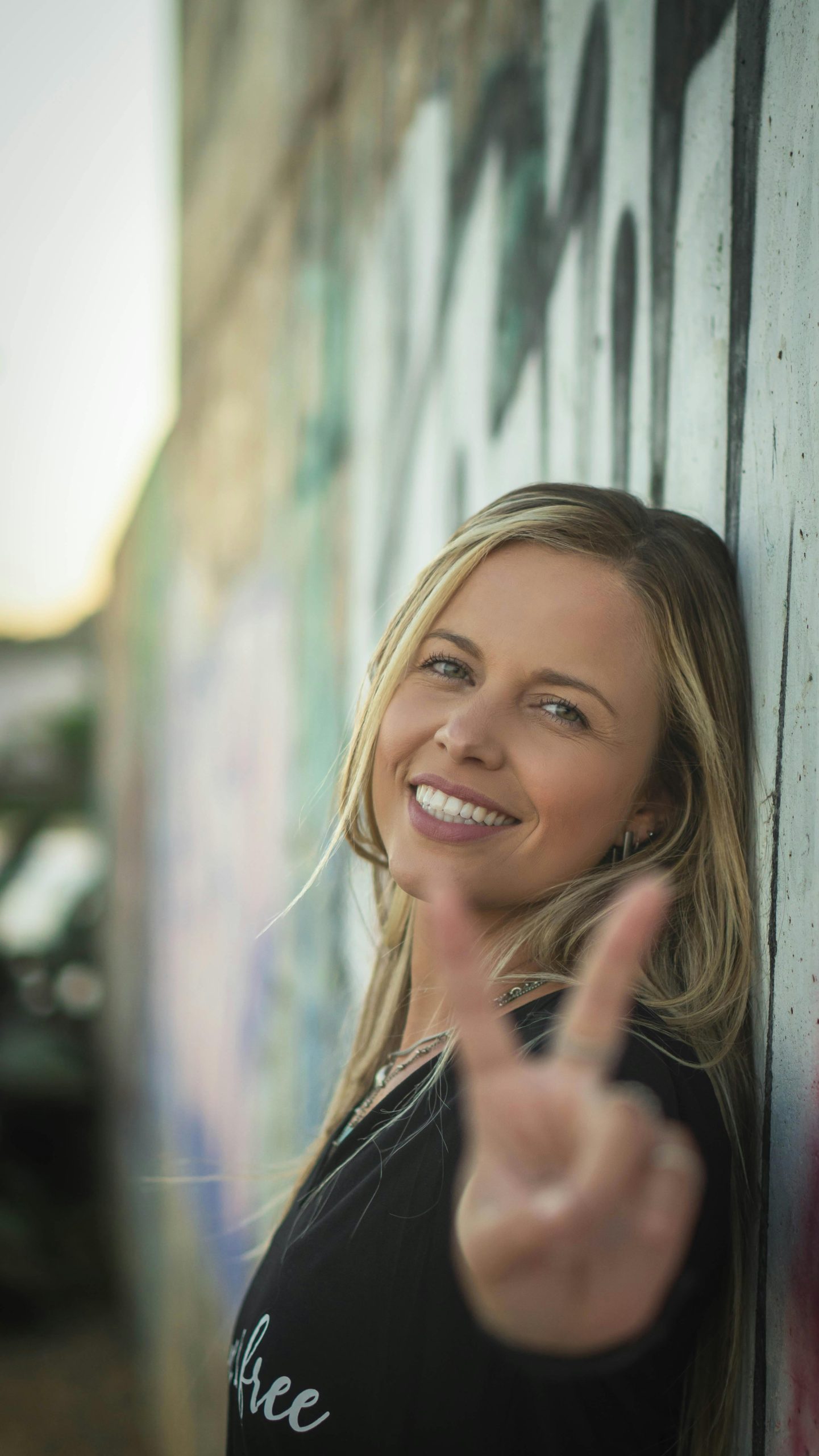 Blonde woman smiling and posing with a peace sign against a graffiti wall outdoors.