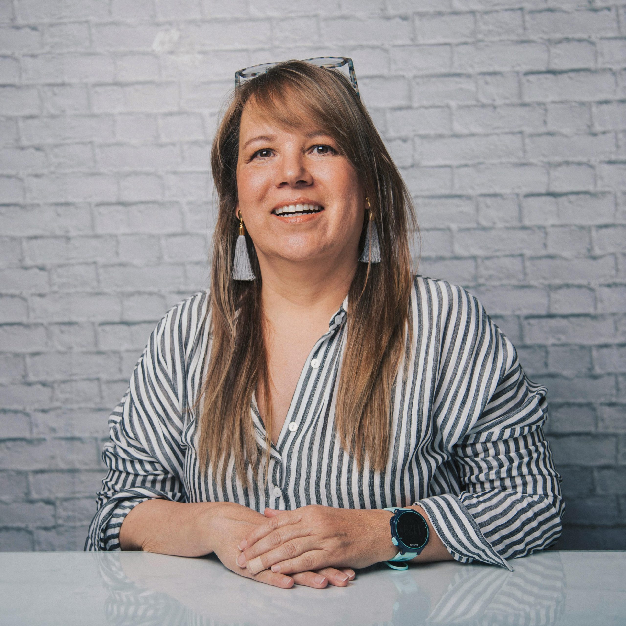 Happy adult female in casual clothes with glasses and earrings looking at camera while sitting at white desk and smiling on white background