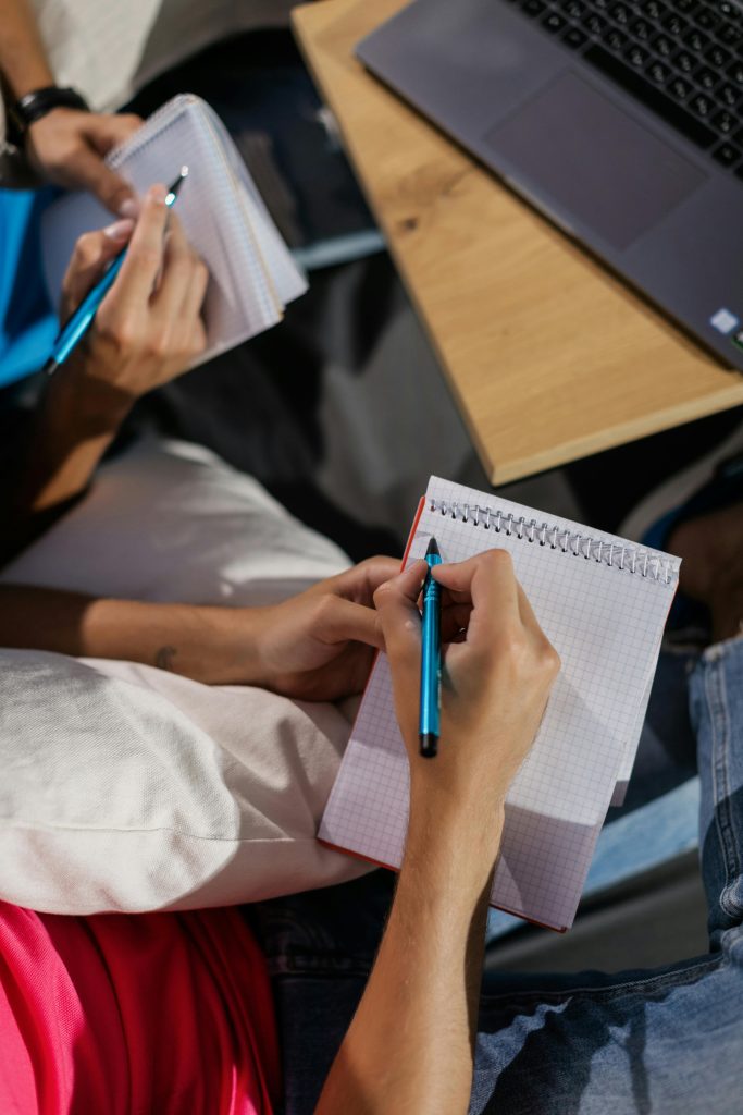 Top view of two adults writing notes in notebooks using blue pens, next to a laptop.