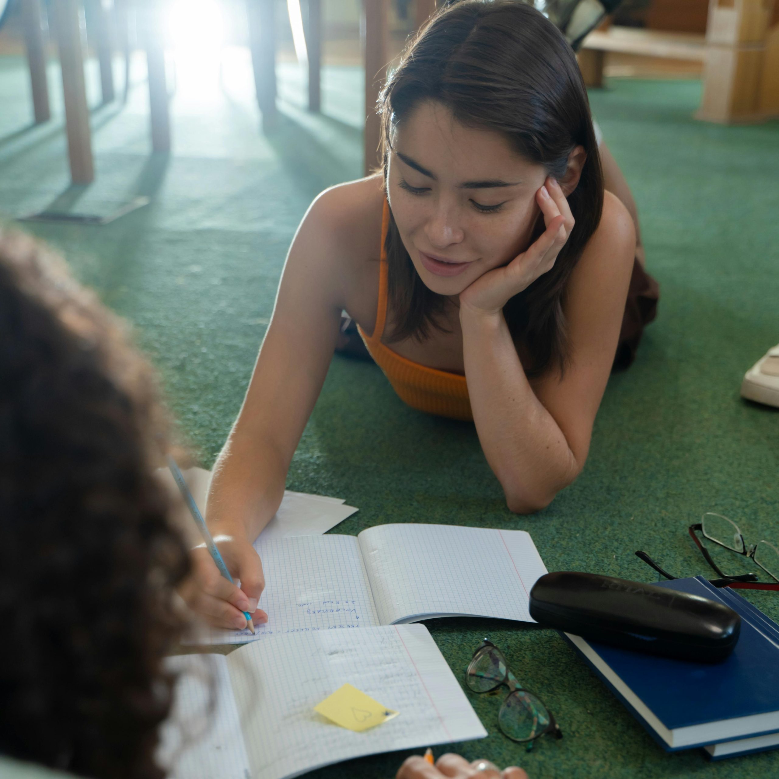 Casual study session: young woman writing notes with a classmate indoors.
